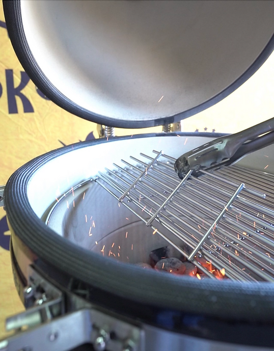 Tongs lifting the hinged cooking grate above glowing charcoal inside an open kamado grill.