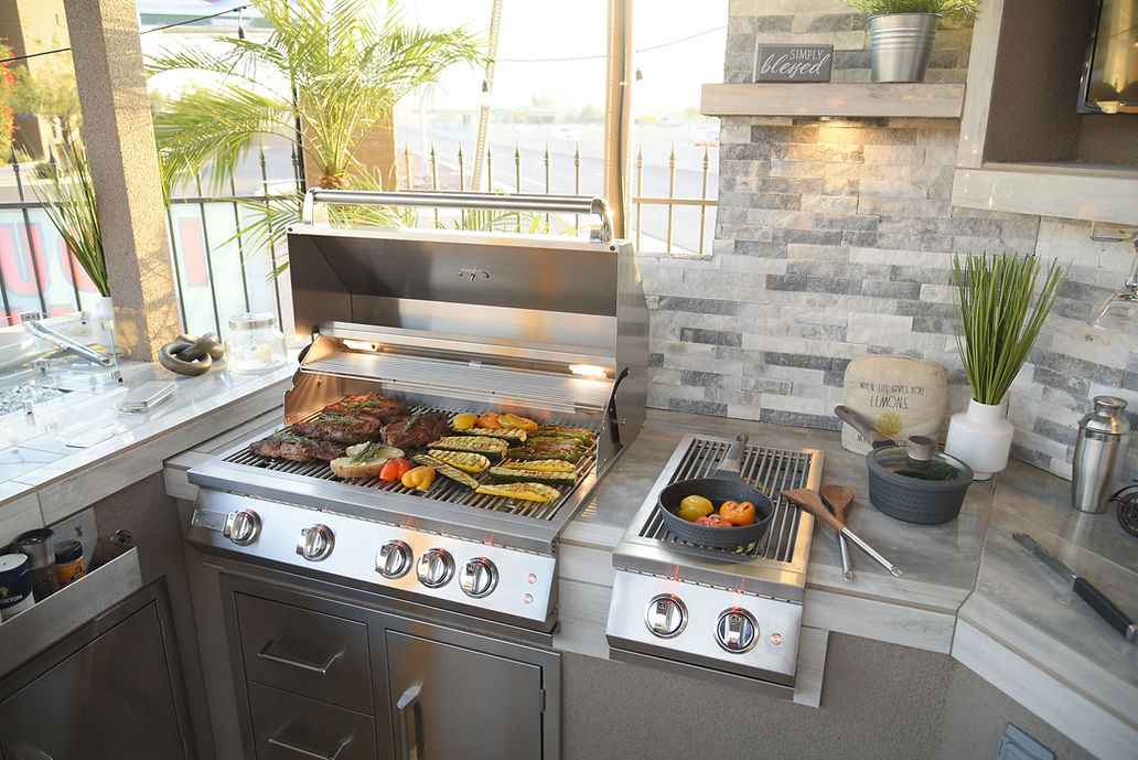Open Kokomo grill cooking meat and vegetables beside a side burner with a skillet.