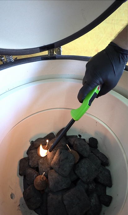 Gloved hand using a long lighter to ignite charcoal inside the ceramic kamado grill.