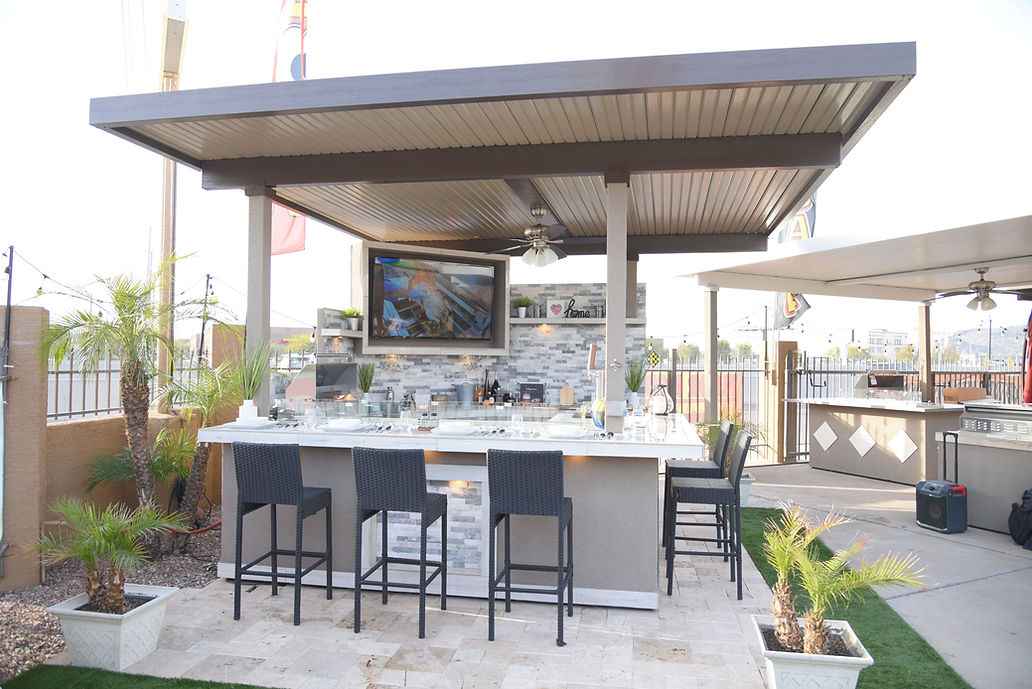 Front view of Kokomo Bermuda covered outdoor kitchen with TV, ceiling fan, bar stools and potted palms.