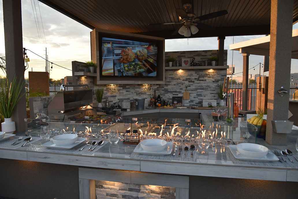 Kokomo Bermuda bar set for dining with lit countertop fire feature, mounted TV and grill behind it.
