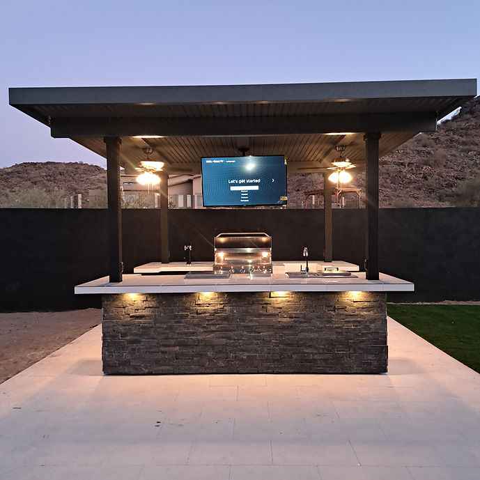 Front view of Kokomo Big Island outdoor kitchen with roof, mounted TV, built-in grill and illuminated stone-look bar.