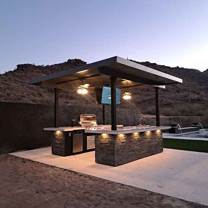 Angled view of Kokomo Big Island outdoor kitchen with covered bar, mounted TV, grill and stainless-steel storage.