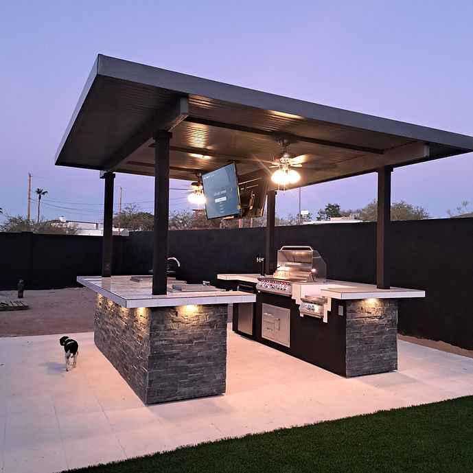 Kokomo Big Island outdoor kitchen showing covered counters, mounted TV, sink, grill and illuminated exterior.