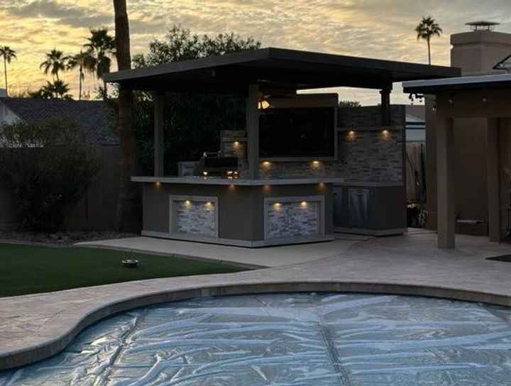 Kokomo Bermuda outdoor kitchen at sunset beside a covered pool, with mounted TV and exterior lighting.