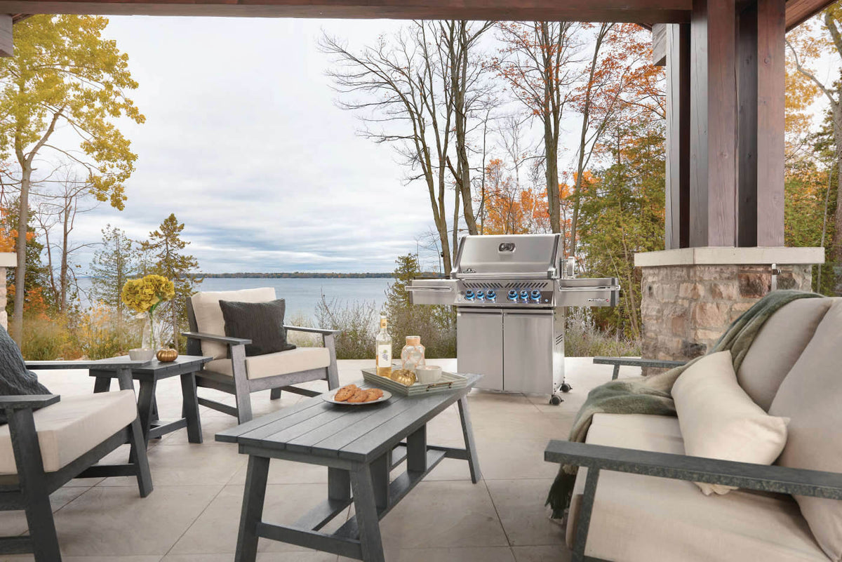 Stainless steel Napoleon grill on a covered patio beside lounge furniture overlooking a lake.