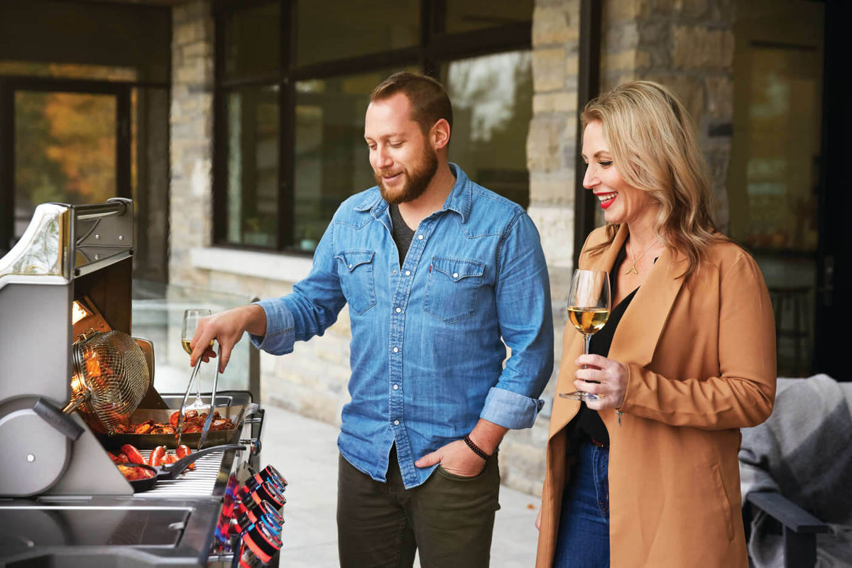 Two people beside an open Napoleon grill while one uses tongs to turn food.