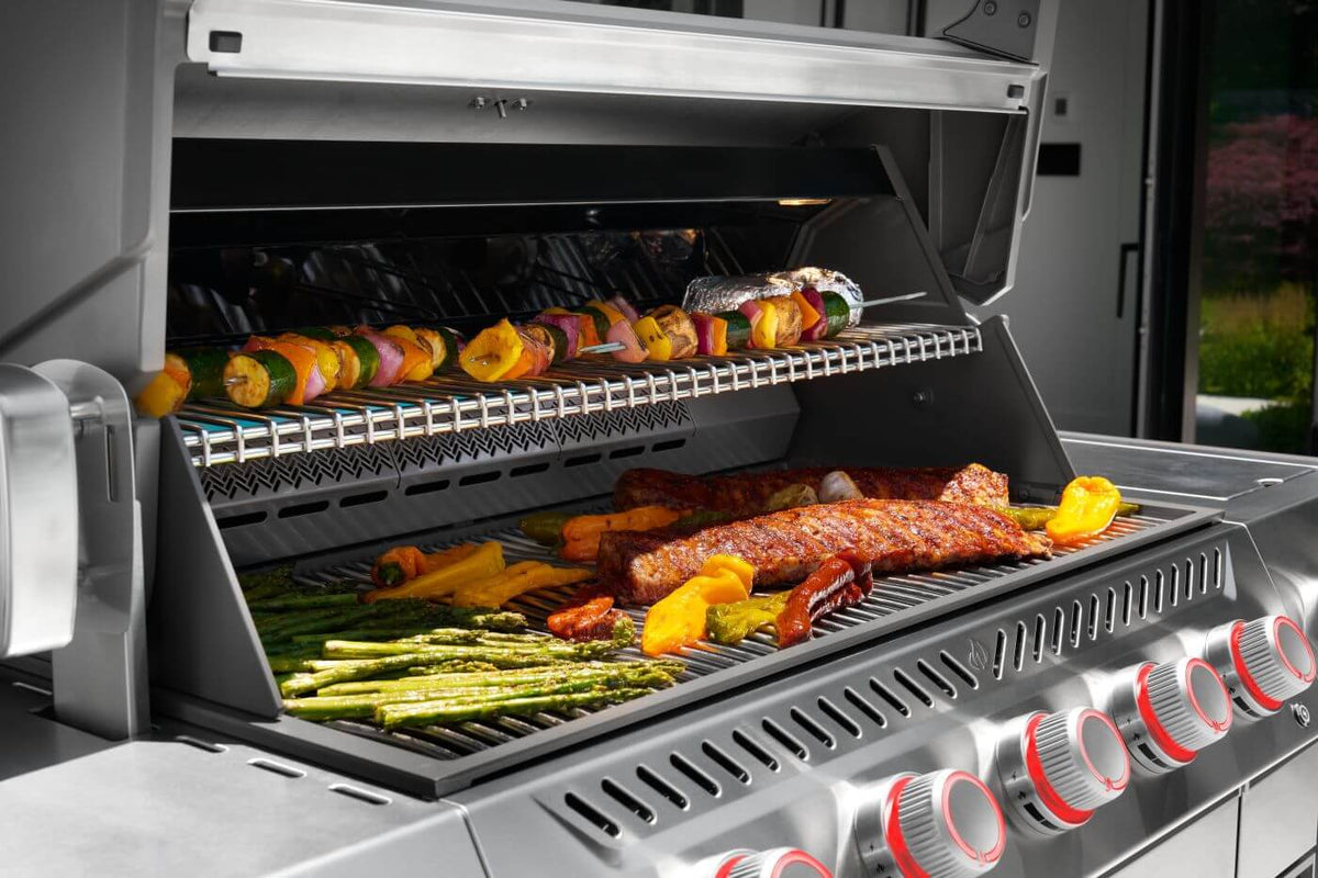 Prestige PRO 665 Elite cooking surface with meat and vegetables on the grates and skewers on the warming rack.