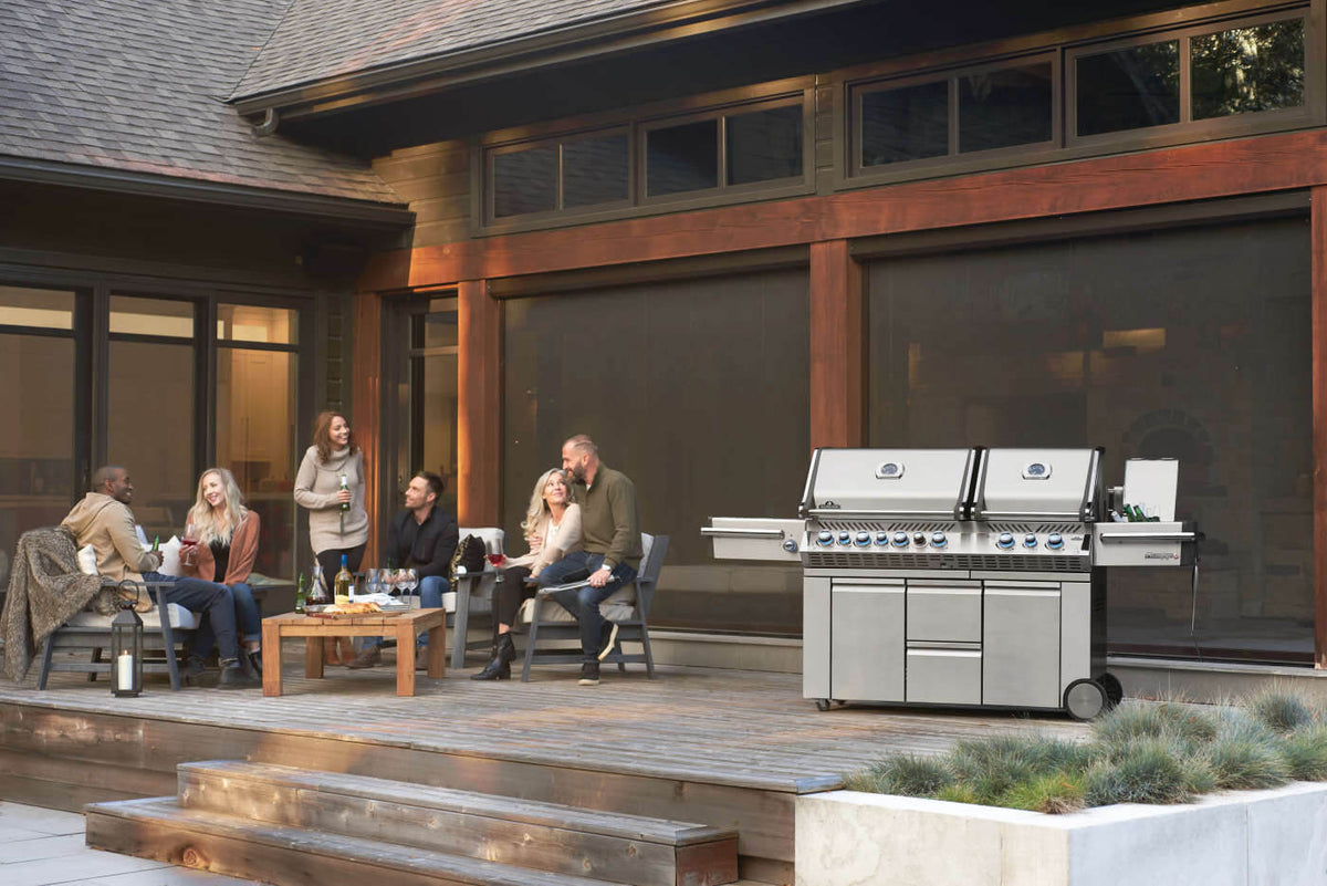 Napoleon Prestige PRO 825 gas grill on a deck beside a group seated around an outdoor table.