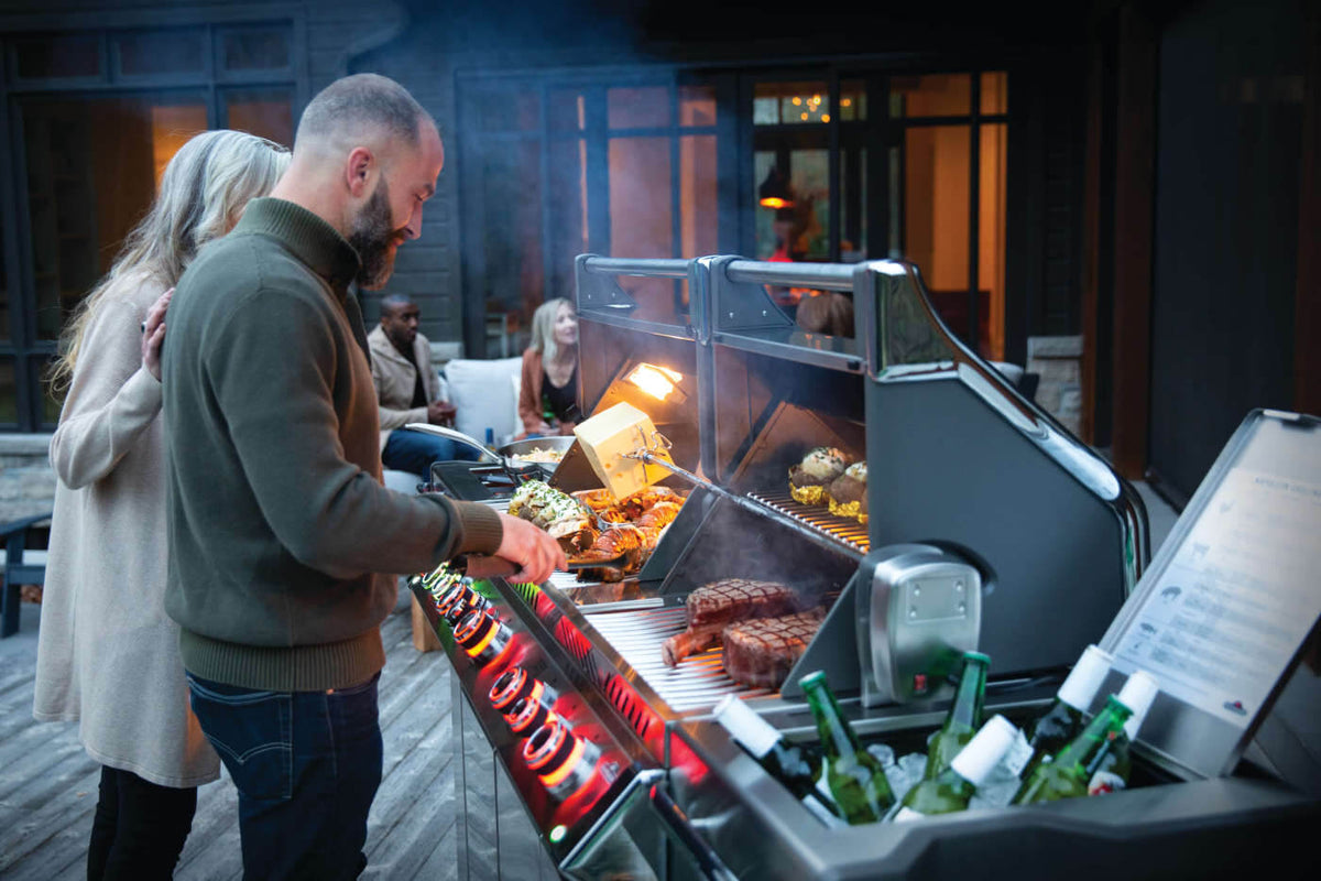 Two people cooking on a Napoleon Prestige PRO 825 gas grill with both lids open and illuminated controls.