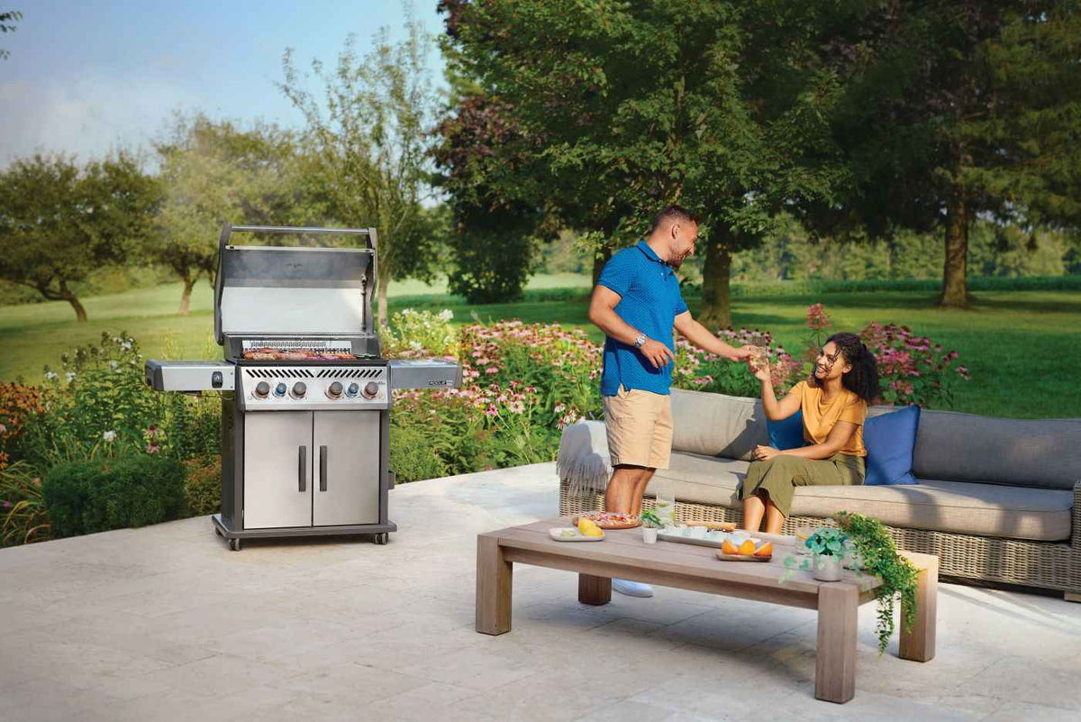 Two people beside a Rogue PRO-S 525 RSIB gas grill with lid open on a garden patio.
