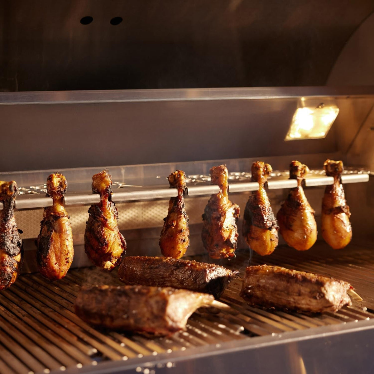 Chicken legs suspended from the Blaze LTE PRO warming rack above steaks on the cooking grates